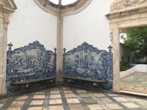 The outdoor room has a marble tiled floor with square pink tiles and grey rectangular border tiles. We are looking into the corner of the room and on walls adjacent to the corner are stone benches surrounded by blue and white Portuguese tile work. The tiles are painted and glazed with medieval scenes. The tiles have a decorative edge and crosses atop the right and left sides of the tile picture. On each outer side of the benches is an open entry surrounded by pink marble. Though the right doorway we can see a tree and shrubs.