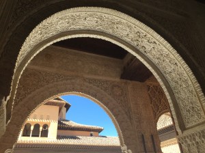 Looking through a Moorish carved stone double arch at the Alhambra. The carvings are intricate, with floral designs bordered by striped graphics. In the background is a tile-roofed building with a dormer and pink walls. The sky is a bright blue contrast.
