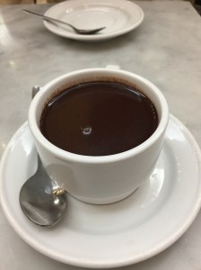 A white coffee cup with saucer and spoon on the left of the cup. The cup is filled with thick hot chocolate. The table is white and grey marble and there is another white plate and spoon in the background.
