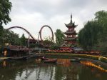 Tivoli Gardens is an amusement park. At the front of the scene is a man-made pond surrounded by shrubs and yellow flowers. There are three rowboats on the pond with a person in each boat. Beyond the pond is a roller coaster with a loop. To the right of the roller coaster is an Asian pagoda. Trees frame the photo on each side.