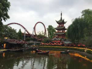 Tivoli Gardens is an amusement park. At the front of the scene is a man-made pond surrounded by shrubs and yellow flowers. There are three rowboats on the pond with a person in each boat. Beyond the pond is a roller coaster with a loop. To the right of the roller coaster is an Asian pagoda. Trees frame the photo on each side.