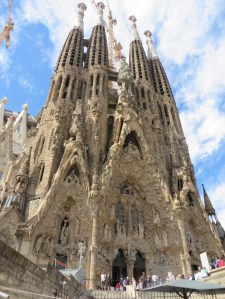 Facade of the Sagrada Familia by Gaudi in Barcelona. The Sagrada Familia cathedral is concrete colored and is ornate in a way that makes it look like a melting cake. There are 4 towers that are rounded cylinders with many openings in vertical columns. There are two doors at the entrance to the cathedral with statues of various biblical figures above the doors.