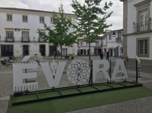 Sign spelling out Evora in town square with white buildings surrounding and trees in the square.
