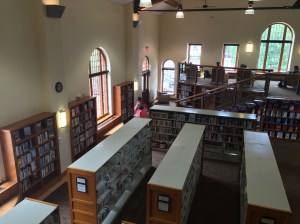 Interior of the Summit County Library South Branch in Breckenridge, Colorado, showing rows of shelves of the book collection, with seating on a raised area at the back of the room. Windows line the walls and the ceiling is very high, about 2 stories above the floor of the library. A female librarian talks with a man standing in the middle of the room.